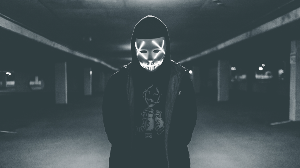 person in dark clothes and a lit up mask stands ominously in a parking garage in black and white