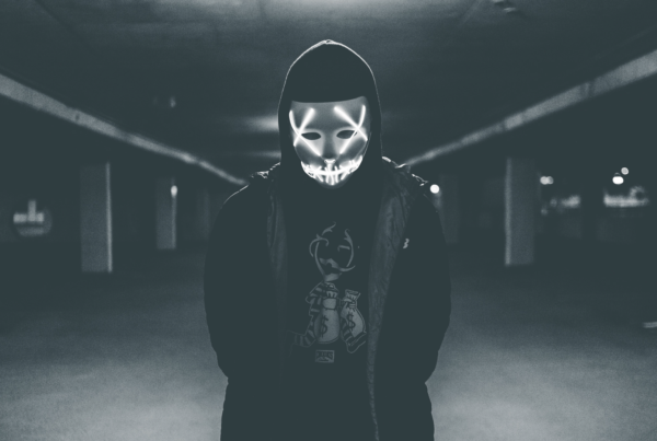 person in dark clothes and a lit up mask stands ominously in a parking garage in black and white